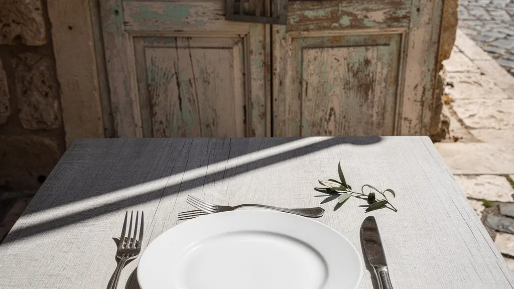 A symbolic still life of an empty white ceramic plate and unused cutlery on a sun-bleached wooden table outside a shuttered Mediterranean restaurant during afternoon siesta hours